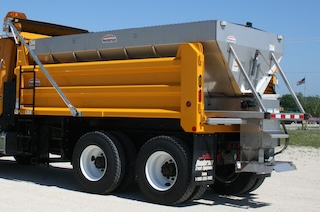 yellow dump truck with a gray metal spreader off the back sitting on concrete on a sunny day